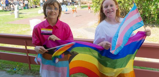 To jenter står med trans og regnbogeflagg.